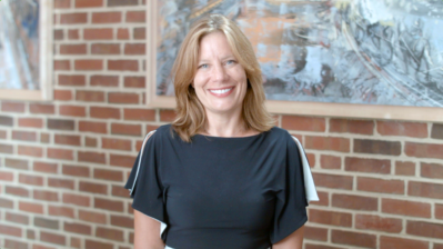 Headshot of a person smiling, standing in front of a brick wall with artwork hung on it. The person is wearing a black top with white accents.