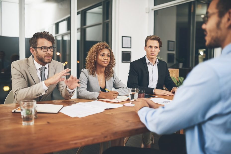  A workplace interview with an interviewer and a candidate sitting across from each other at a desk, with another person sitting at the head of the desk, all showing enthusiasm and interest.