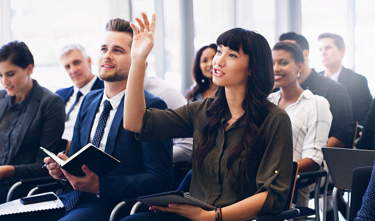 A small group of people in a seminar or training session. A person in the foreground raises a hand, indicating engagement or a question. Others hold notebooks and appear attentive.