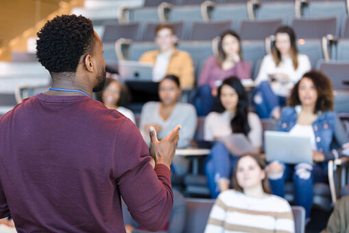 An instructor is addressing a small group of students in a lecture hall. The students are seated, some with laptops and notepads, engaged in the session.