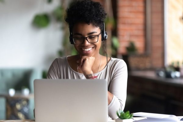 A person wearing headphones and glasses is smiling while using a laptop. The setting appears to be a cozy indoor workspace with plants and soft lighting.